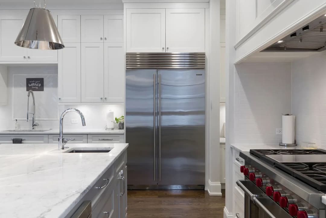 Modern white kitchen featuring stainless steel refrigerator, marble countertop, and gas stove.
