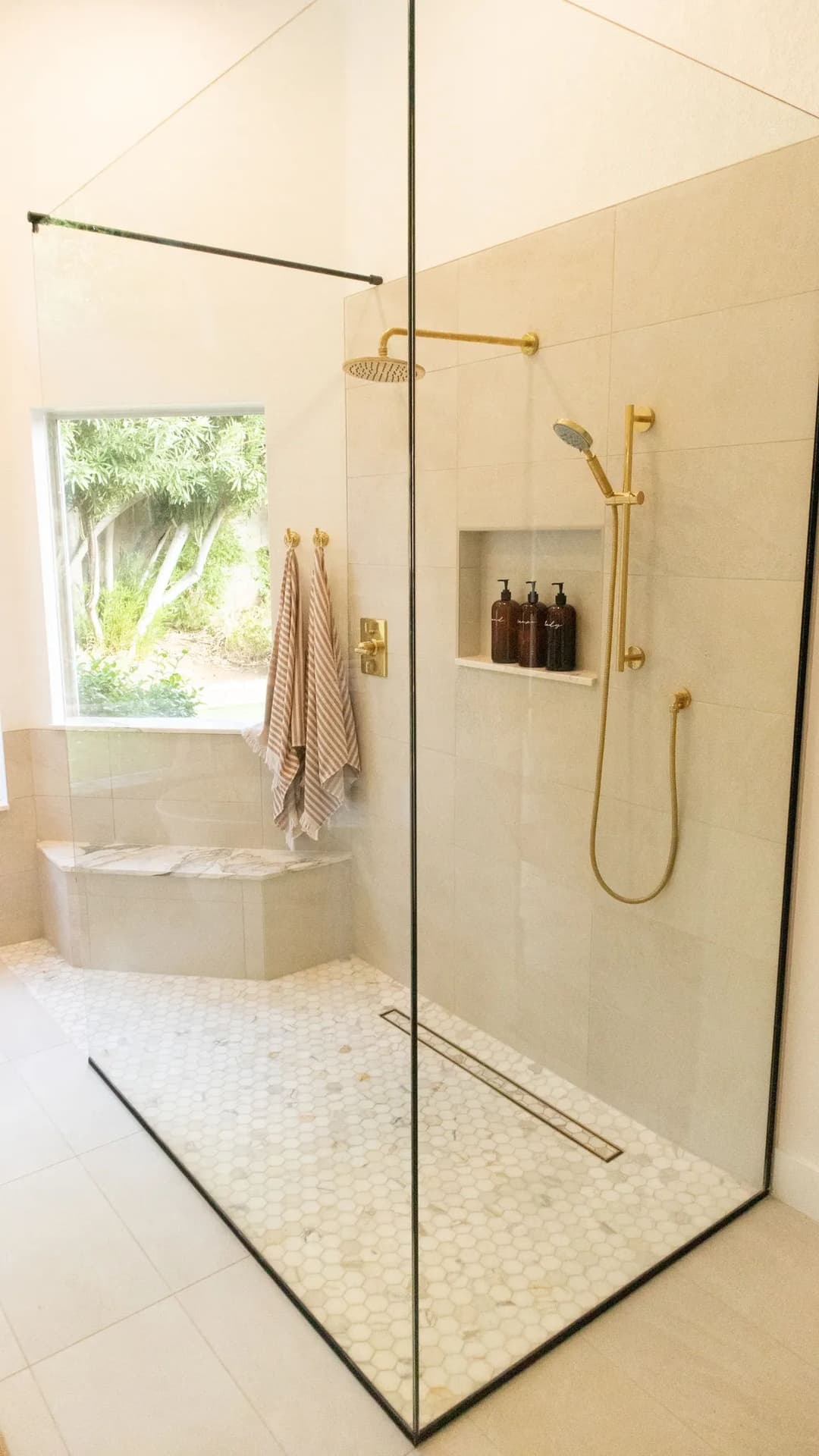 Modern glass shower with gold fixtures, stone seating, and natural light from a window.