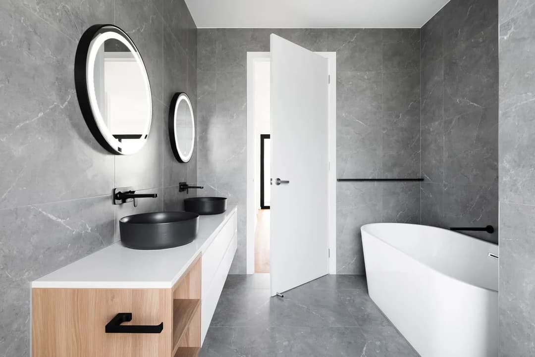 Modern bathroom with gray marble walls, circular mirrors, black sinks, and a freestanding tub.