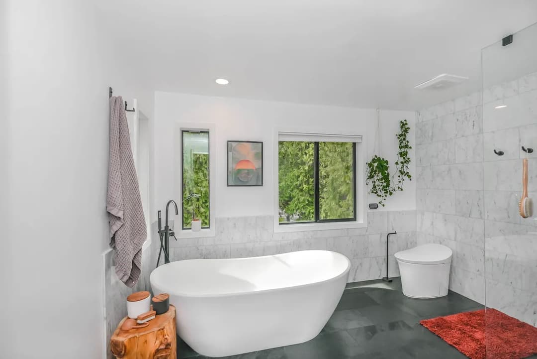 Modern bathroom with freestanding tub, large window, and marble accents. Bright and airy design.