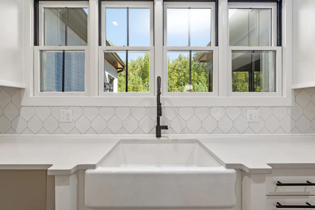 Farmhouse sink under window with modern faucet and white tiled backsplash in bright kitchen.