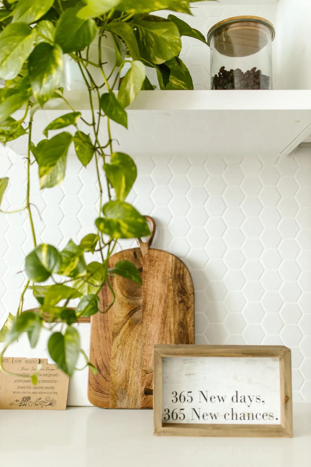 Wooden cutting board and motivational sign surrounded by greenery on a kitchen shelf.