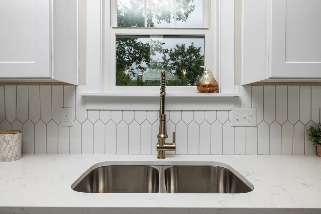Modern kitchen sink with gold faucet, white cabinets, and hexagonal tile backsplash.