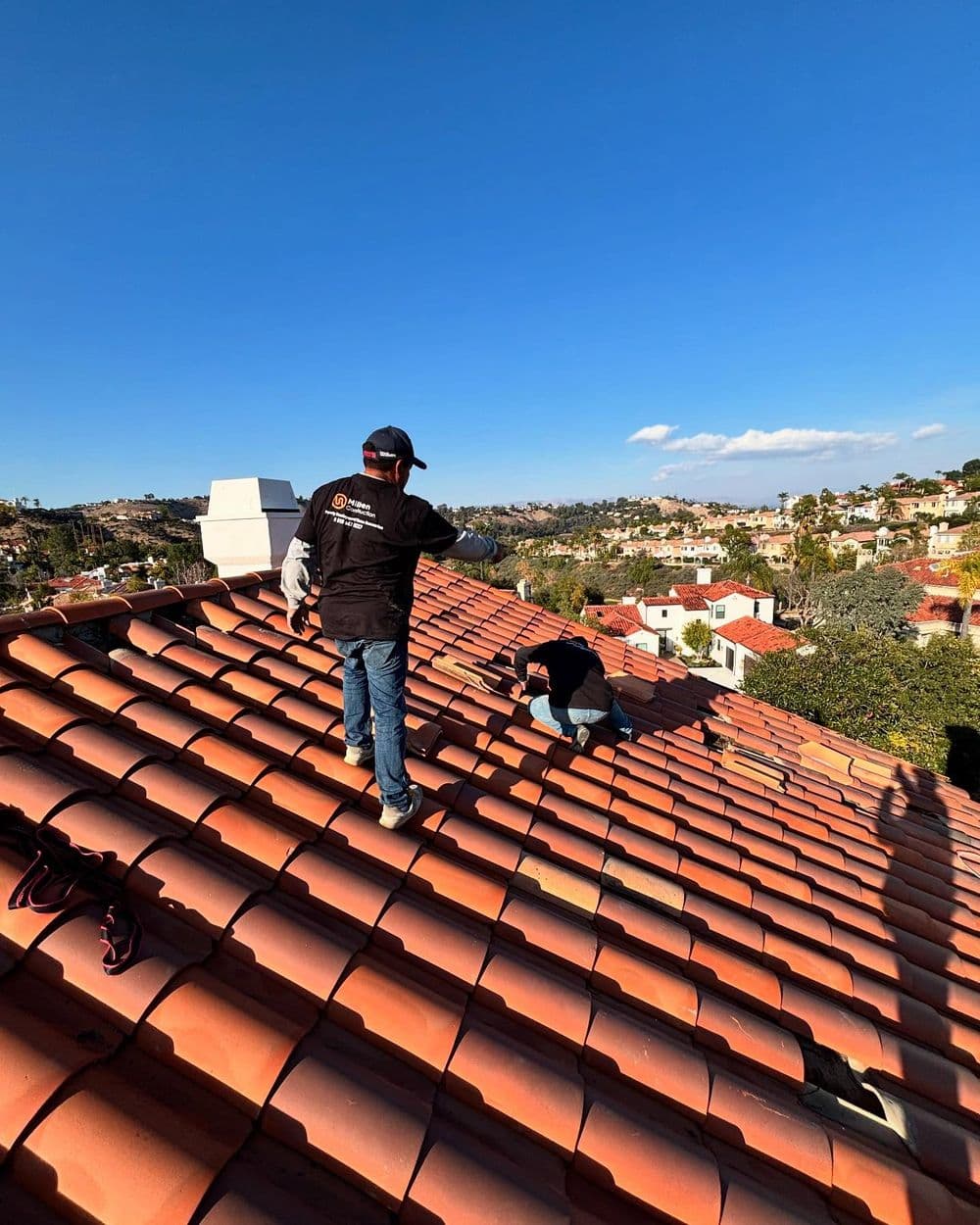 Roofing professionals working on a tiled roof with a scenic view of homes below.