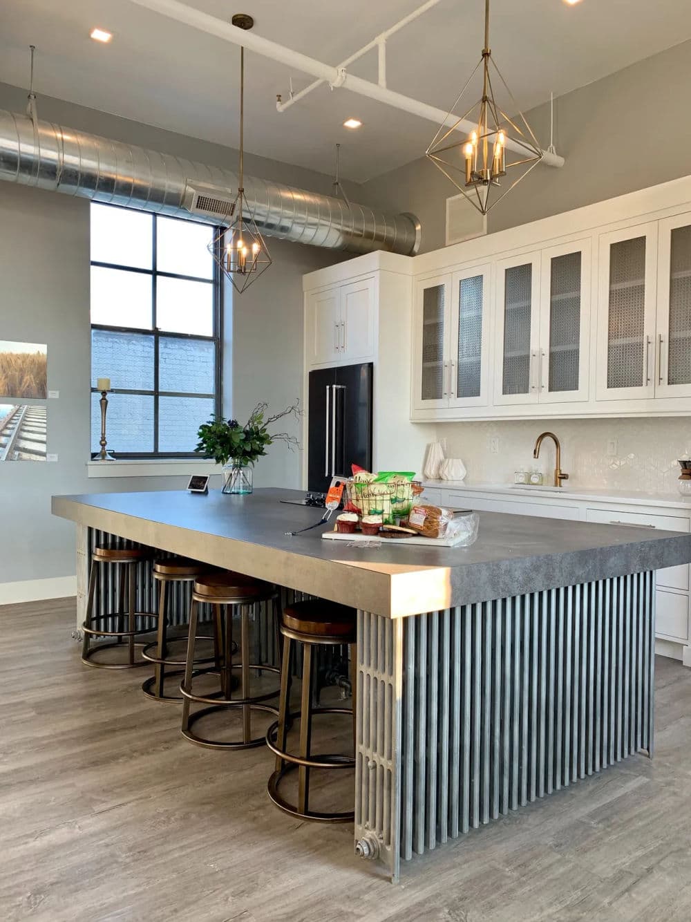 Modern kitchen with gray concrete island, stools, stainless steel accents, and large windows.