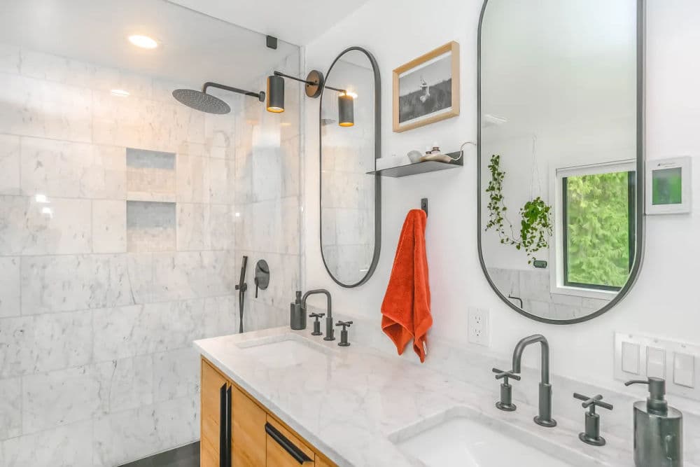 Modern bathroom with marble tiles, dual mirrors, stylish faucets, and a vibrant orange towel.
